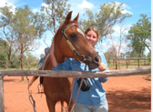 Picture of lady standing beside her saddled horse giving it a hug around the neck .  The background is blue sky, red soil and eucalyptus trees.